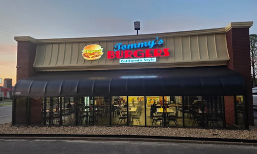 Exterior of Tommy's Burgers restaurant at dusk, with illuminated signage showing a burger. Glass windows reveal empty tables and chairs inside.
